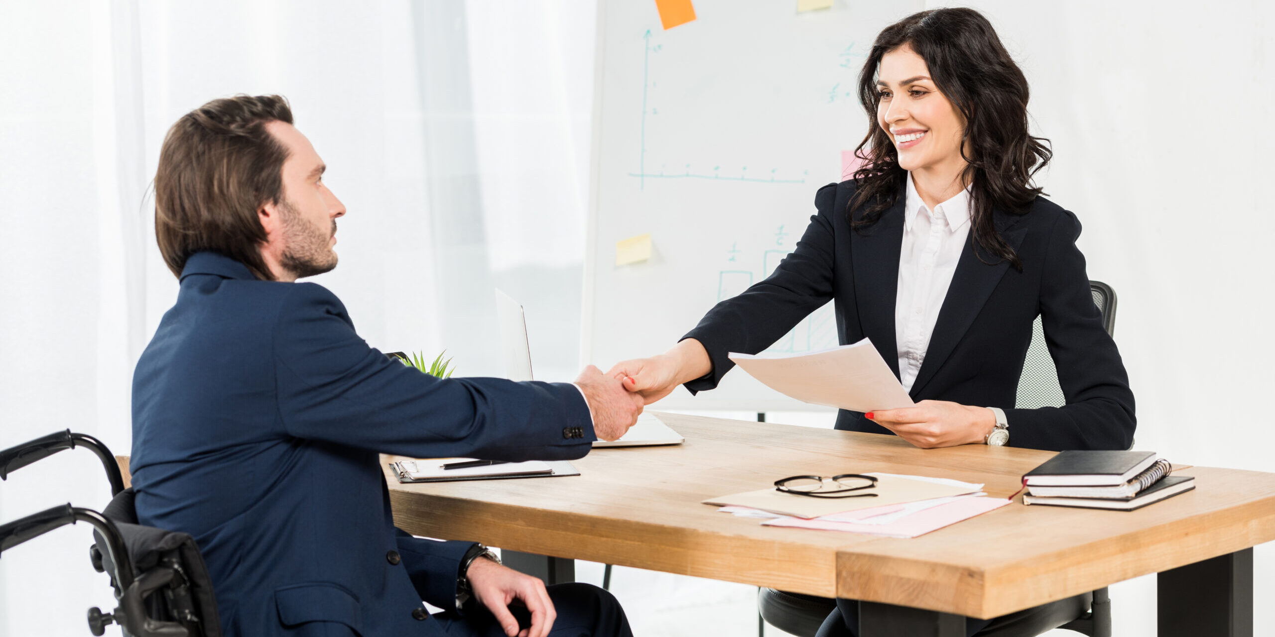 happy recruiter shaking hands with disabled man while holding documents in office