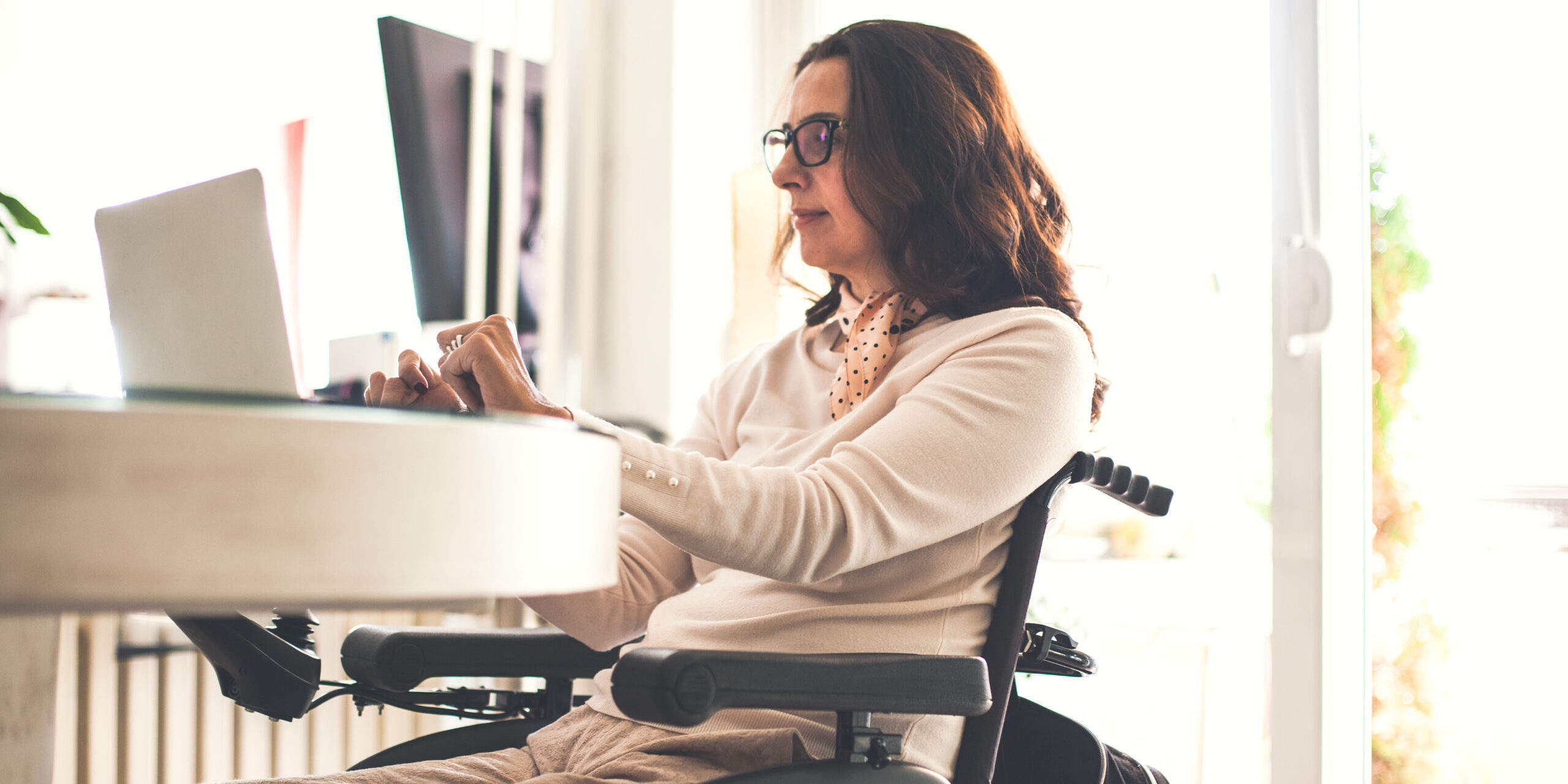 Woman in wheelchair is working in the comfort of her home. She is using laptop at the dining table.