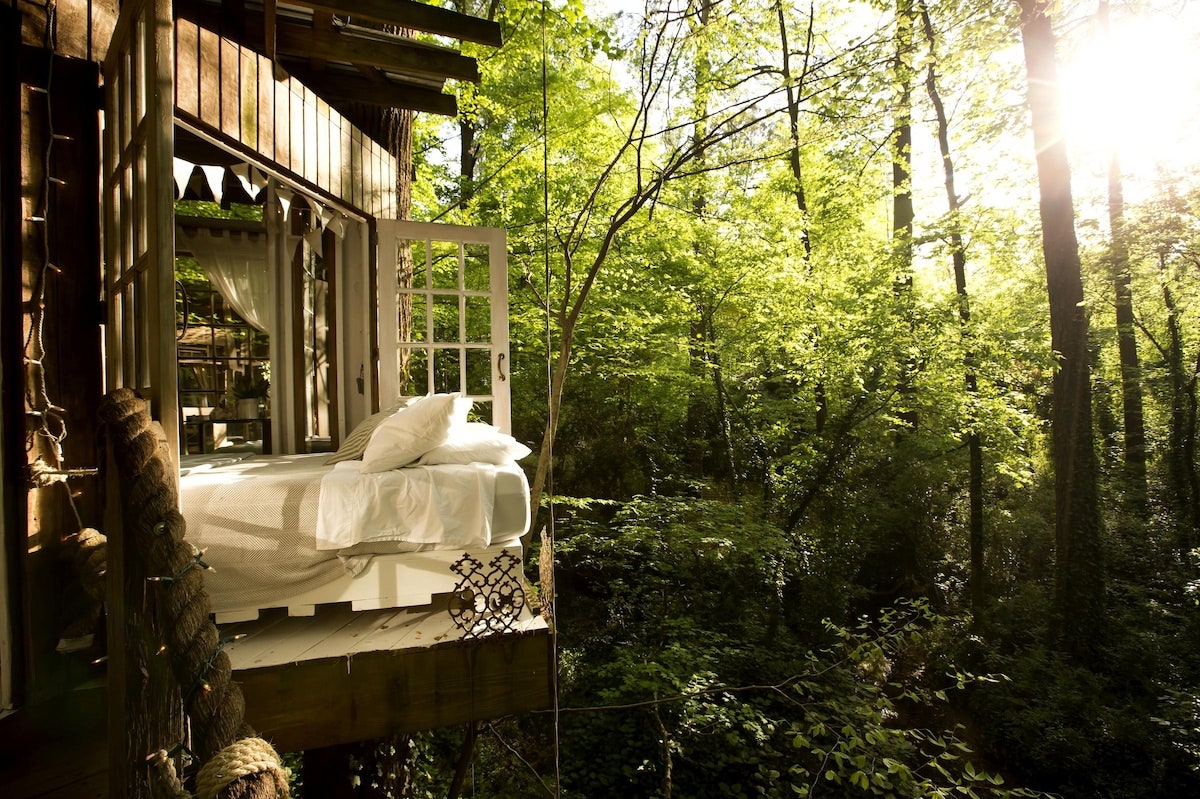 Bed on an open air deck with a forest surrounding it.