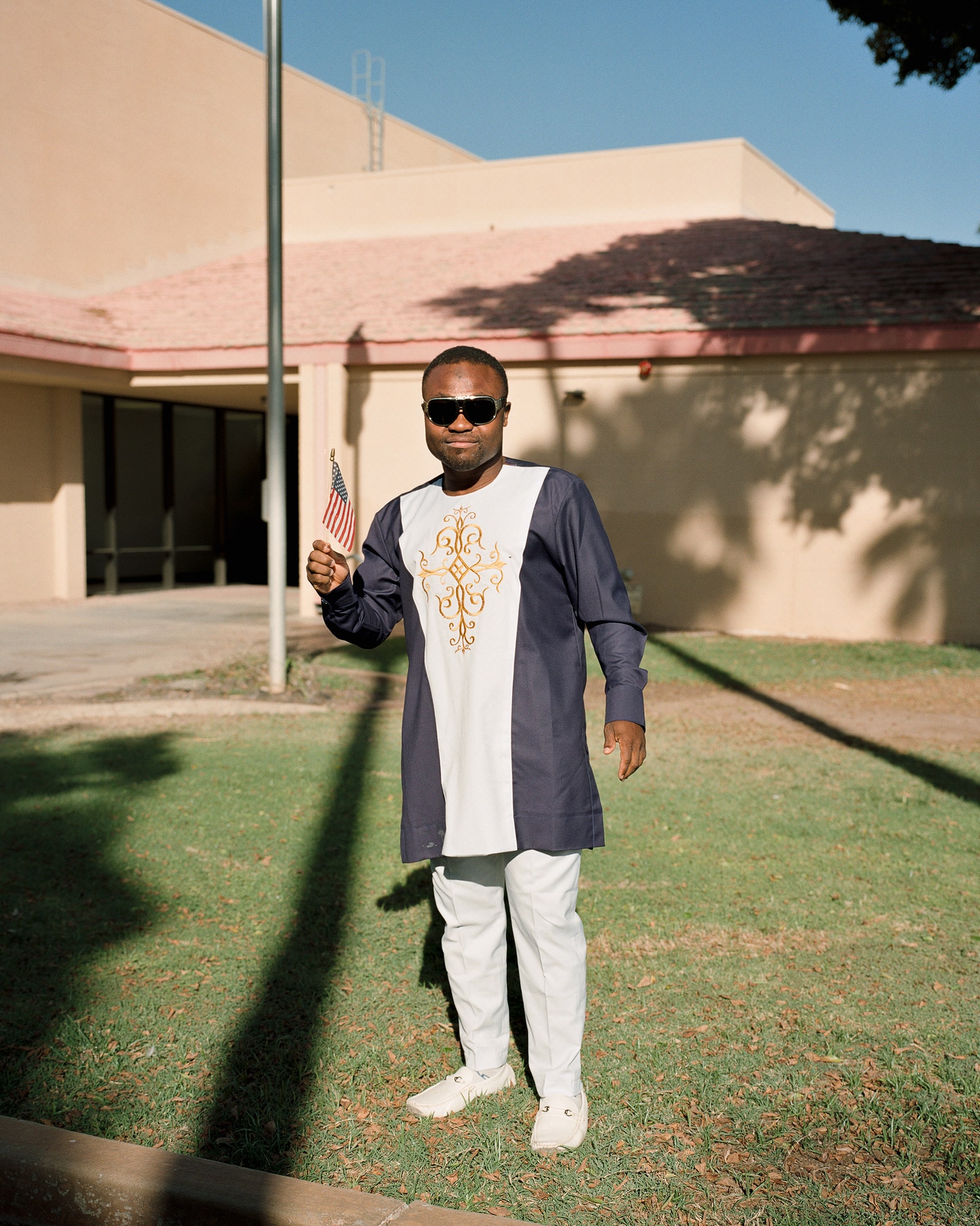 A person standing with a small American flag in their hand. They wear white shoes white pants and a long shirt.