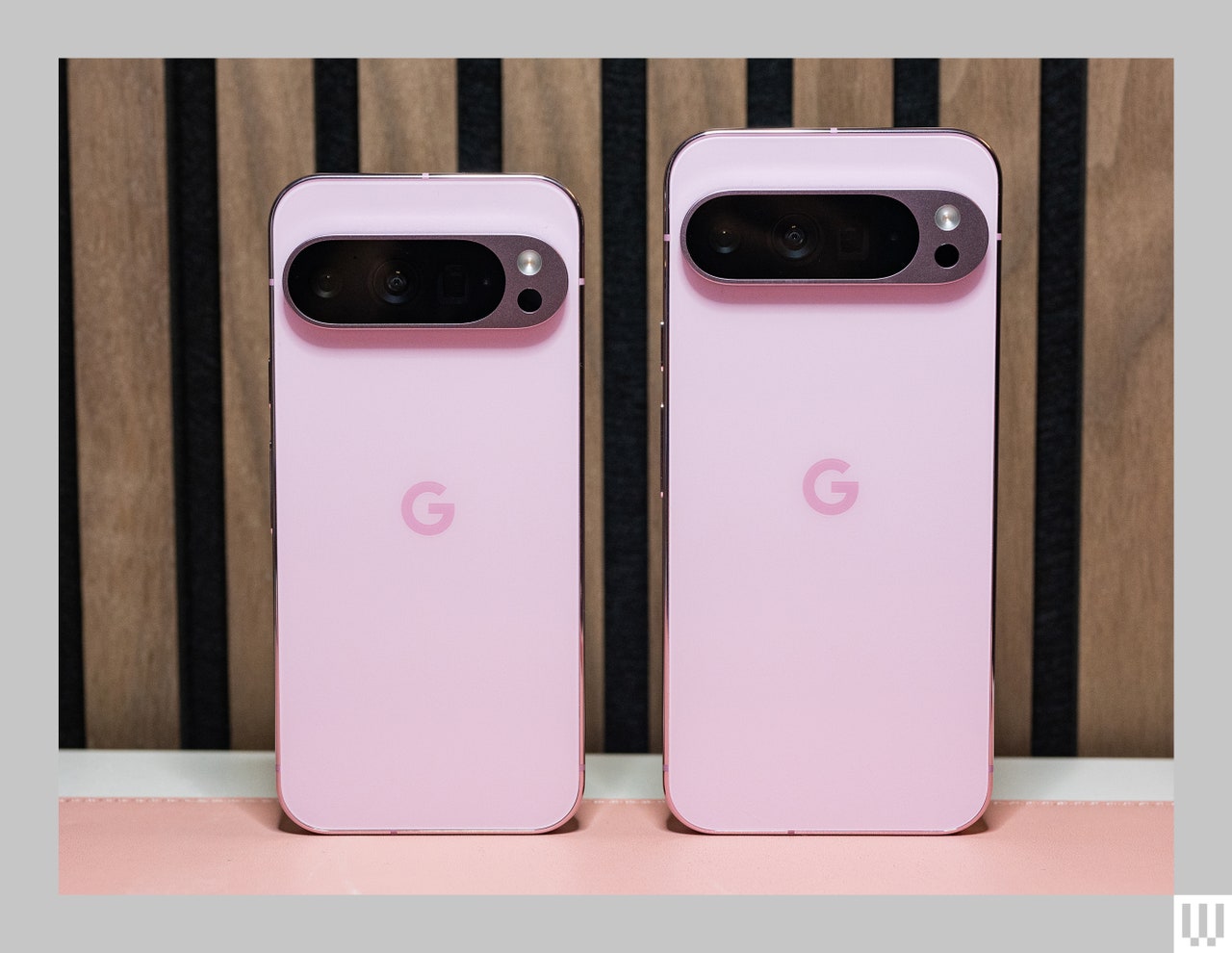 Back view of 2 pink mobile phones, side-by-side showing the oval-shaped cameras, both propped up against wooden panel wall