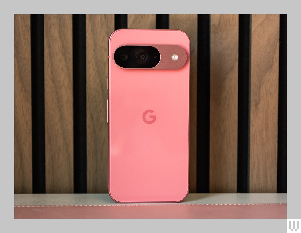 Back view of a pink mobile phone, showing the oval-shaped camera, propped up against wooden panel wall