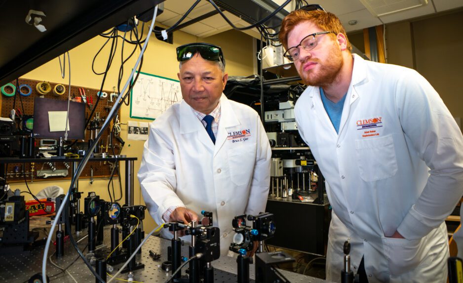 Two male researchers in white lab coats with the Clemson paw on them work at a large computer-like machine with various cords, buttons and controls.