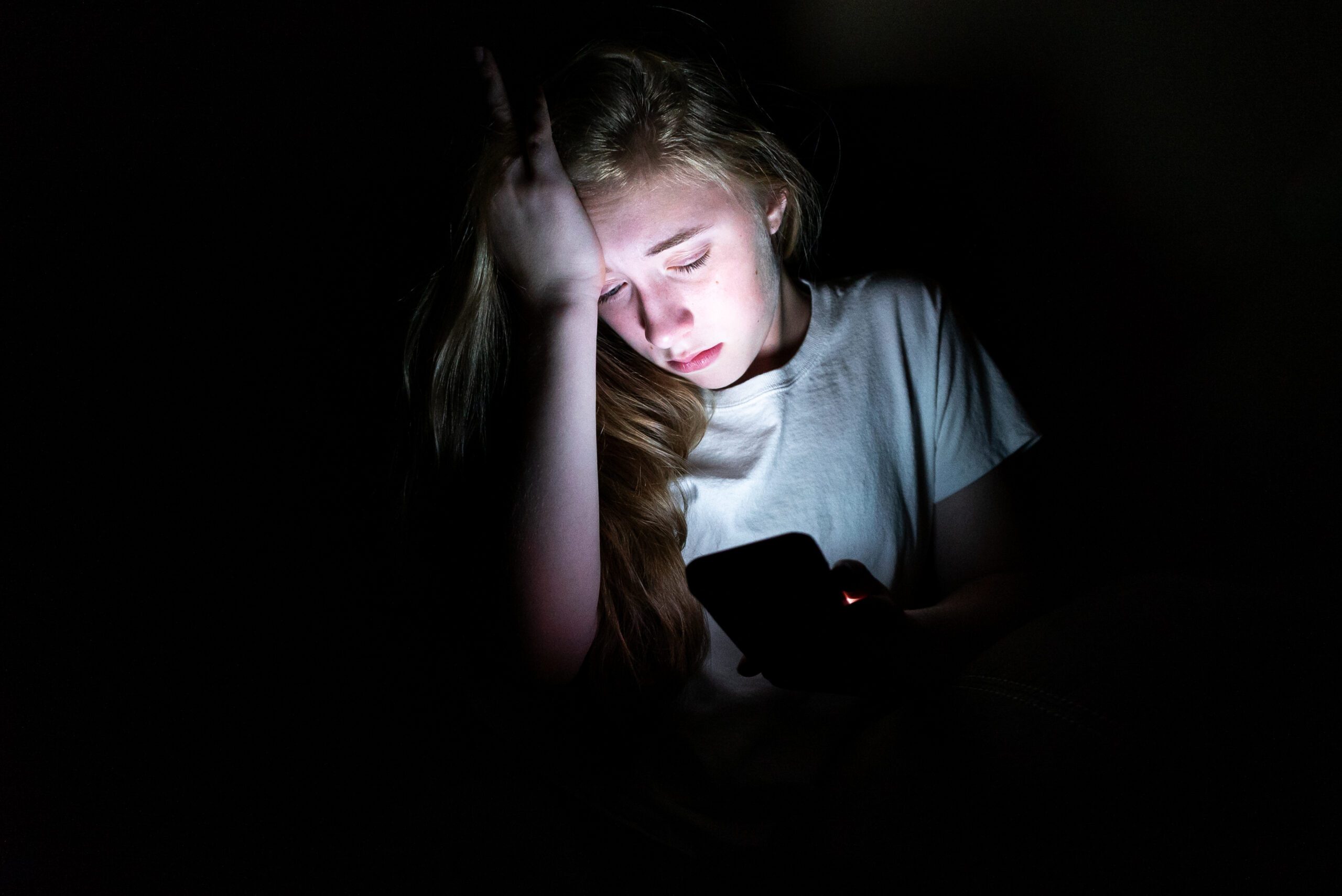 Upset girl sitting in the dark while using her smartphone. The light from the screen is illuminating her face.