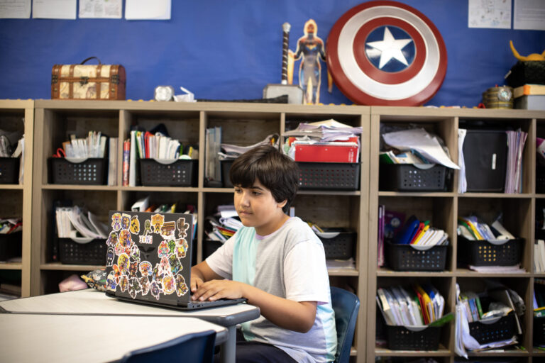 A child working on a laptop covered in stickers, seated in a classroom with shelves filled with books and various decorations in the background.