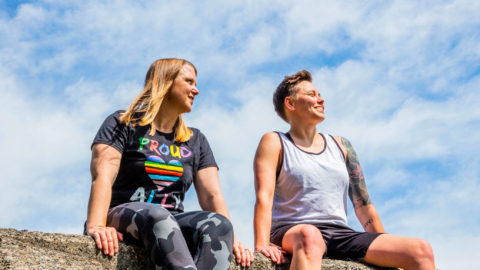 Two women sit on a stone structure at a playground. They are smiling and it's a sunny day.