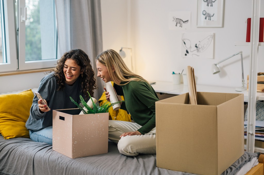 College female teenagers sitting on a bed, using a mobile phone in a dormitory, with boxes around