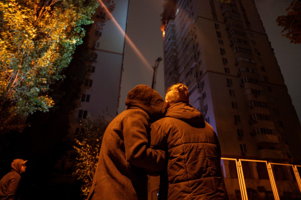 A couple seen from behind embracing each other, watching a fire burn in an apartment building in Kyiv in October following a Russian drone attack.