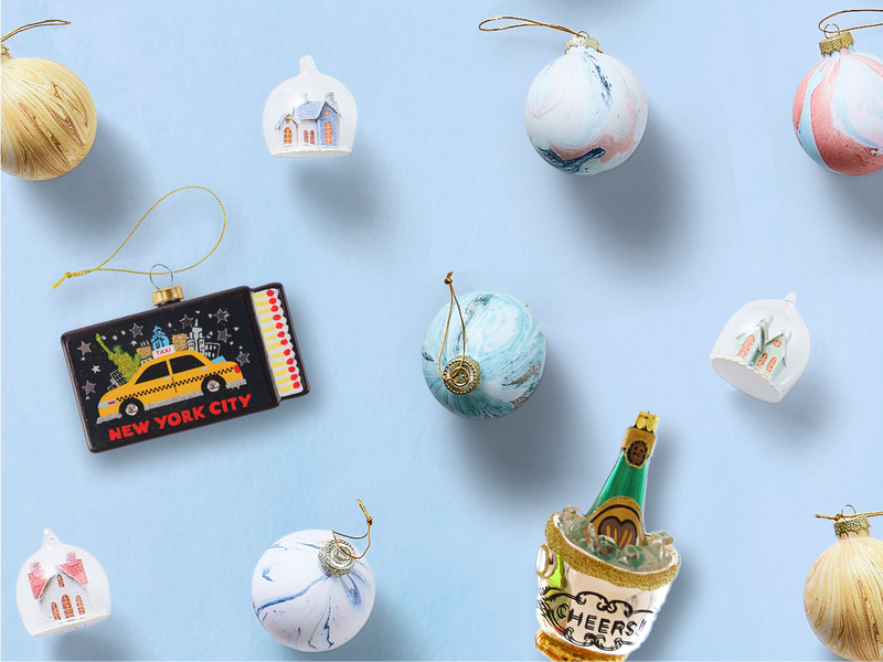 Eleven ornaments are placed sporadically against a baby blue backdrop, featuring a NYC taxi, a champagne bucket, painted baubles, and frosty houses.