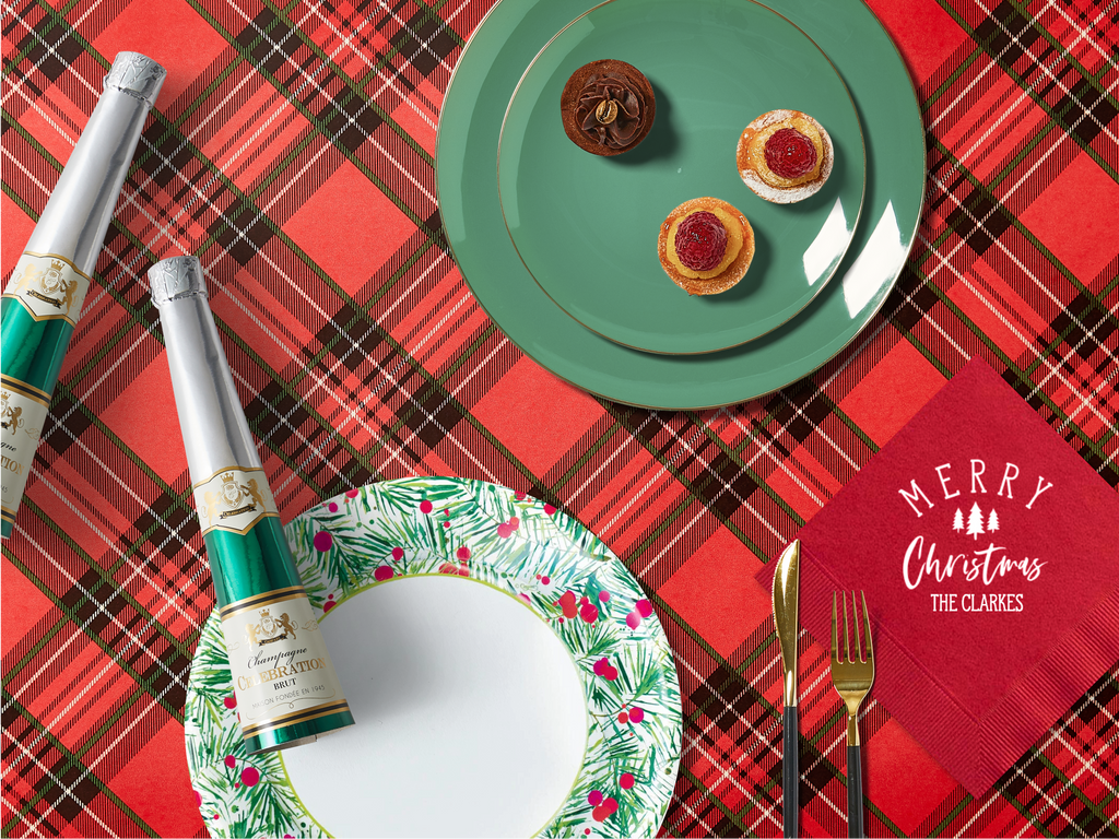 A festive table with a red plaid tablecloth, a green plate with small desserts, holiday crackers, a holly-patterned plate, and a personalized red napkin reading 