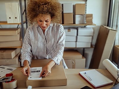 Eine Unternehmerin in einem gestreiften grauen Hemd klebt einen Versandaufnäher auf ein Paket