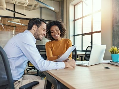 Ein Mann und eine Frau sitzen in einem Büro. Sie zeigt ihm Dokumente und hat den Laptop neben sich geöffnet