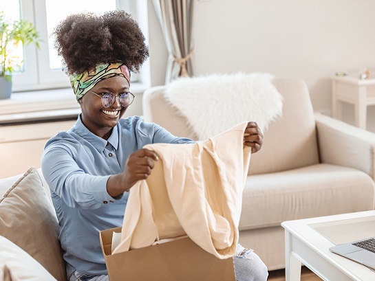 Frau mit vielen Locken und Brille sitzt auf der Couch und packt ihr Paket aus und freut sich