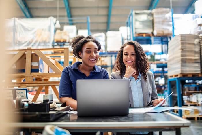 Zwei Frauen sind im Warenlager, stehen vor einem Laptop und schauen sich Paketstatistiken im GLS Kundenportal an