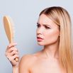 Concept of hair loss. Close up portrait of unhappy sad stressed young woman with long dry blonde hair, she is looking at the brush in hand, isolated on grey background