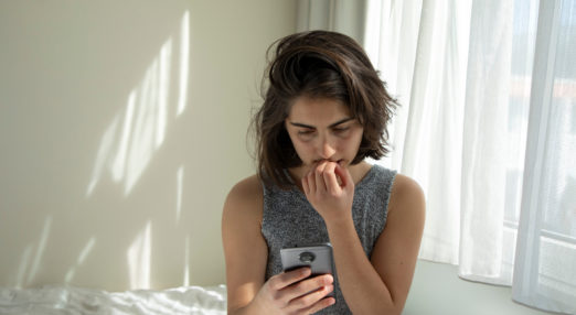 Young brunette woman holding a phone while sat on a bed.