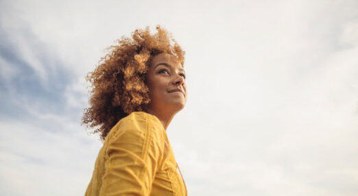 A women, pictured from below, looks into the distance. The sky is blue and she wear yellow and positive expression.