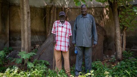 À gauche : Prudencio Legge Lodu, chef spirituel de la communauté Bari, à Juba au Soudan du Sud. Il pose devant Pita, pierre sacrée, dont il a la responsabilité spirituelle. C'est lui qui conduit les cérémonies et rituels. À sa droite : le chef bari Lino Loro Gore, le 21 juin 2024