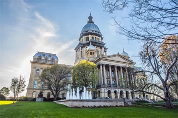 Illinois state capitol building surrounded by grass, trees, and a partially cloudy sky above.