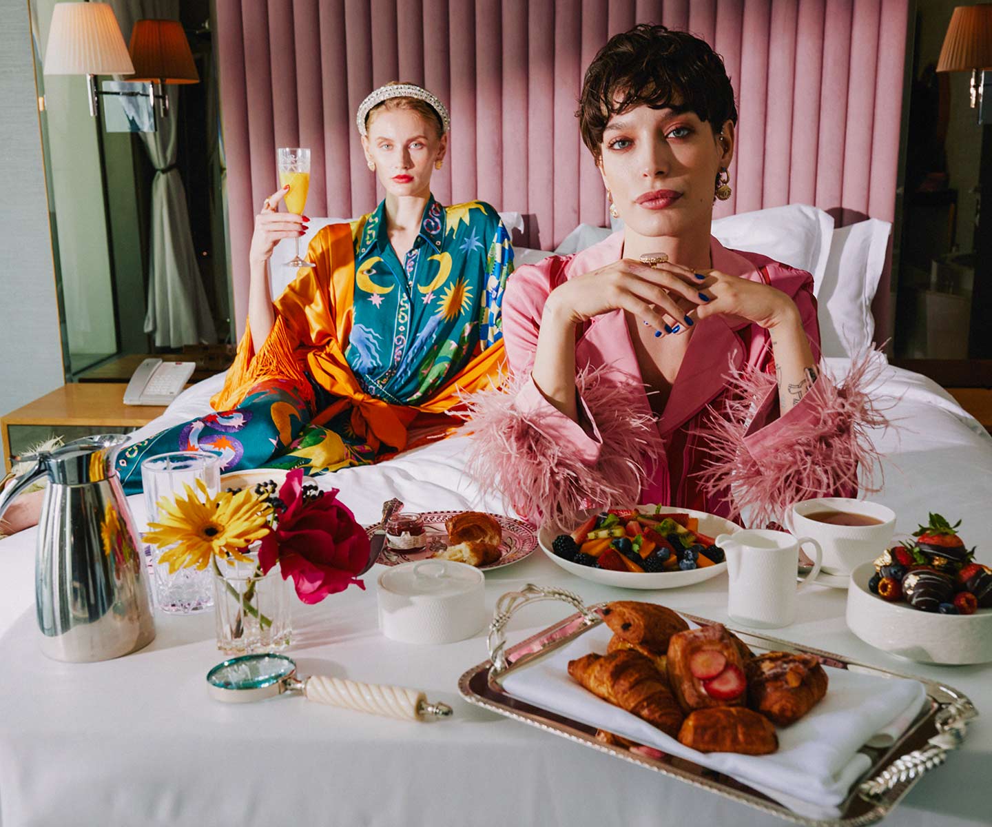 Two elegant ladies enjoying a delightful morning on a luxurious bed, savoring a scrumptious breakfast spread.