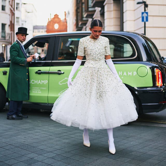 A person walking in a street past other people and a green-and-black car. The person is wearing a bedazzled white top and a tulle skirt, long white gloves, white stockings and white heels.