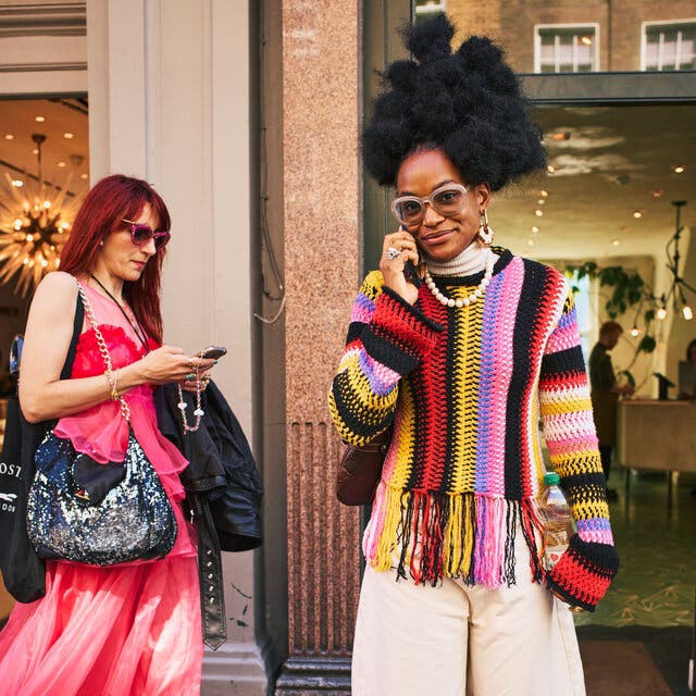 A person standing on a sidewalk. The person has natural hair styled in a sort of Afro and is wearing a vibrant striped sweater with pale wide-leg pants.