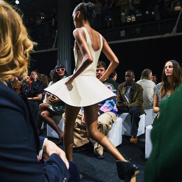 A model, seen from behind, walking on a runway in a pale dress with a wide, short skirt.