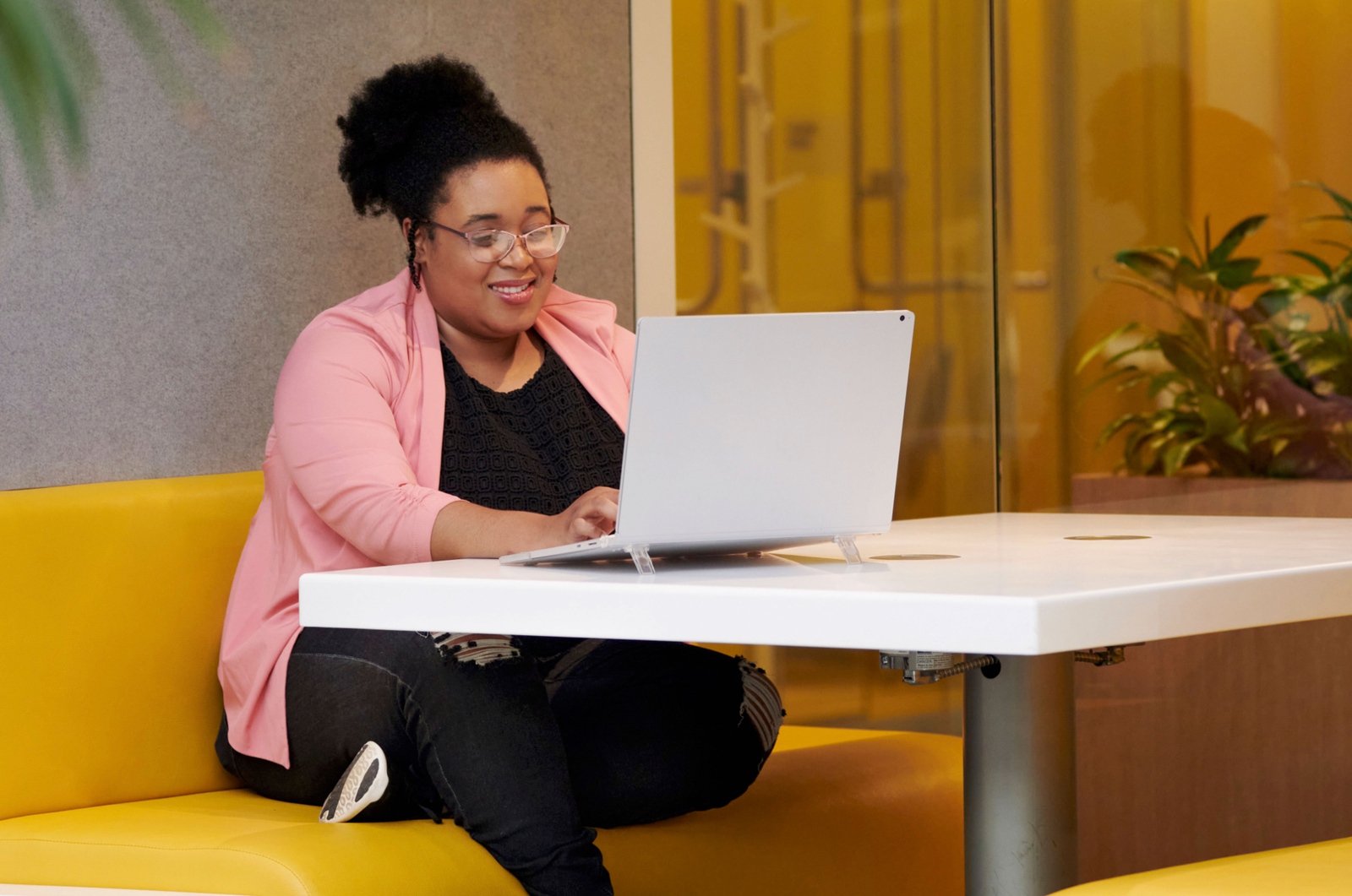 A woman in a pink jacket, perched on a yellow couch works at her laptop surrounded by lush plants.