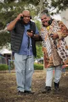 A woman and a man standing in front of trees, listening to music from a device