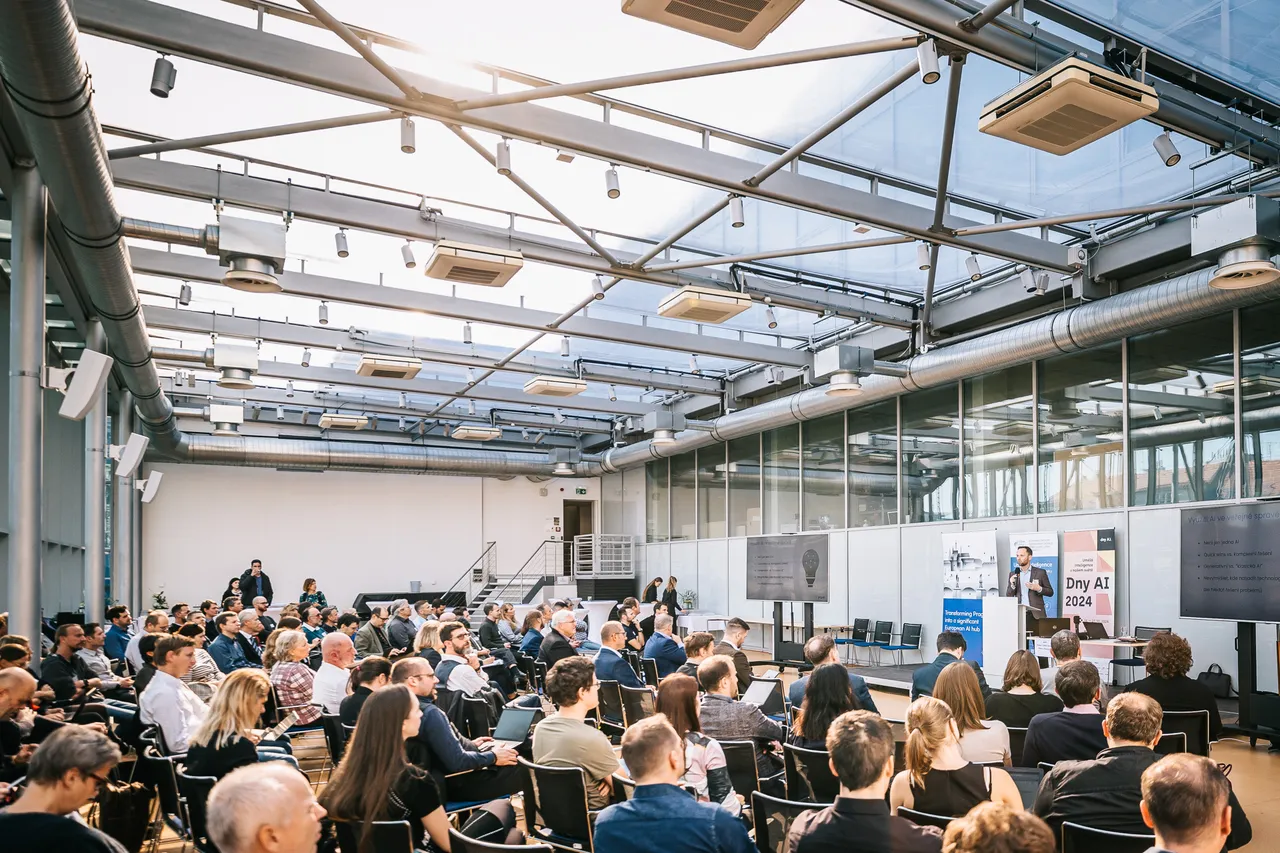 A photo of a crowd in a big room on chairs facing the stage with a speak on the stage.