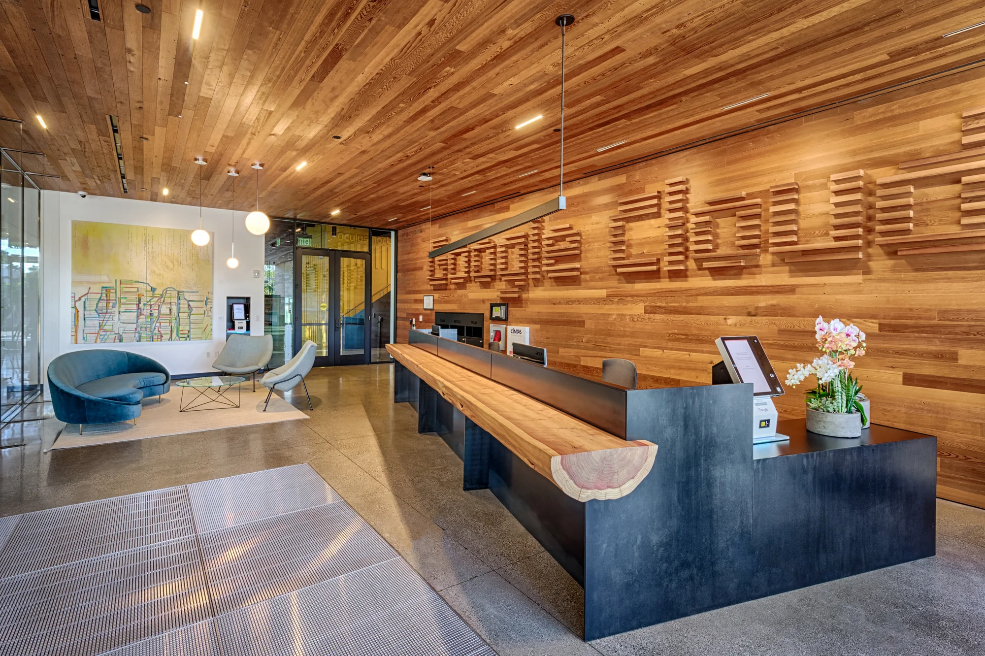 The office lobby has wood paneling on the ceiling and one wall that says 'Google Cloud' with a long reception desk made out of reclaimed redwood. In the corner a couch and two chairs are set against the backdrop of a large piece of abstract artwork.