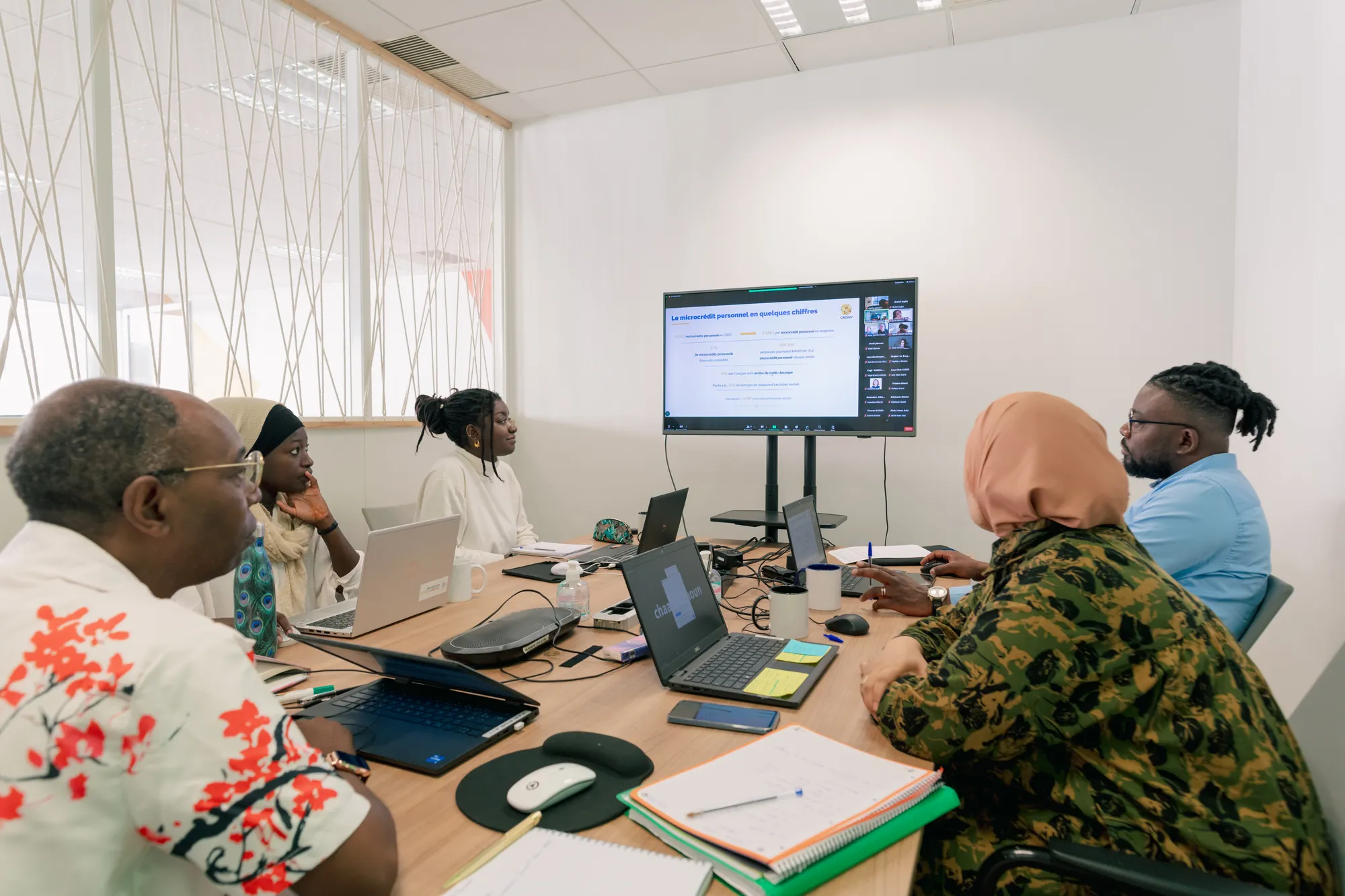 A photograph of a group of people around a desk, all looking at a big screen.