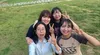 Four women are smiling at the camera while taking a selfie in a grassy field. One woman in a blue shirt is holding her left hand up, while the other three women are making a peace sign with their left hands.