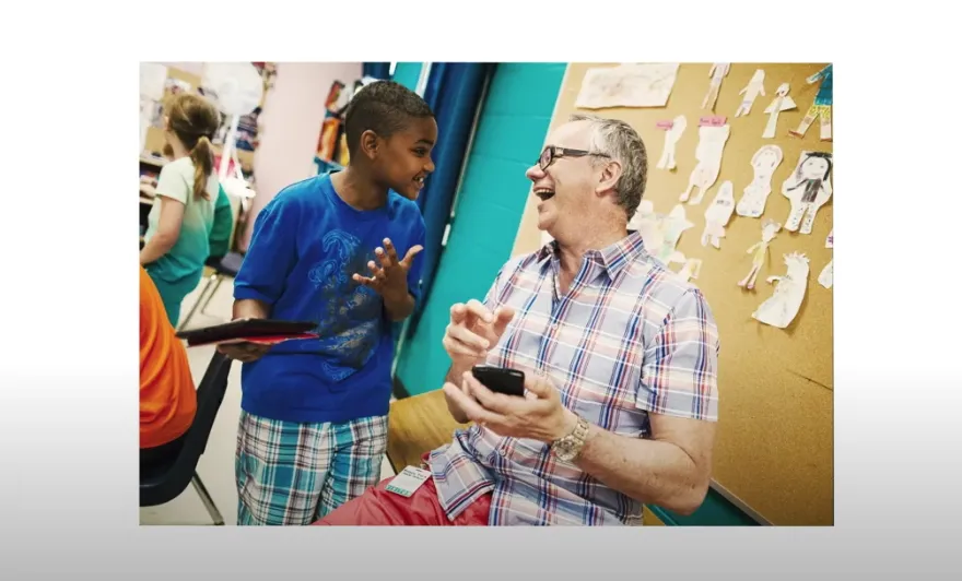 A student holding a tablet talks enthusiastically with a smiling teacher who is holding a phone