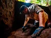 A gloved man excavating in a square hole of earth.