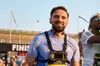 A photo of Fatmir Seremeti, a visually impaired runner, smiling broadly as he has crossed the finish line banner that reads "FINISH". He is wearing a blue running shirt and black harness with a phone on his chest.