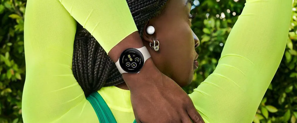 A photo showing a woman wearing a neon yellow long sleeve top from the shoulders up. She is facing away from the camera with her face in profile and stretching her arms behind her head, revealing a Google Pixel Watch 3 on her wrist.