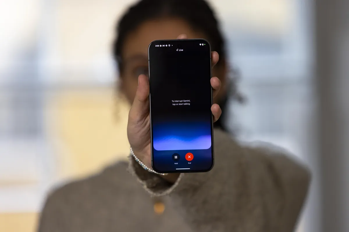 Photograph of a woman holding a phone which shows Gemini Live on the screen (a blue screen with a pause and stop/x button on it.