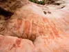 A flat stone wall of the mountainside with rock art paintings in red of large, rectangular bodied creatures in proportion to their raised arms and legs.