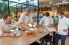 Four chefs gather around a table with two pizza pies in the center. One chef gestures towards the pies while the others review recipes.