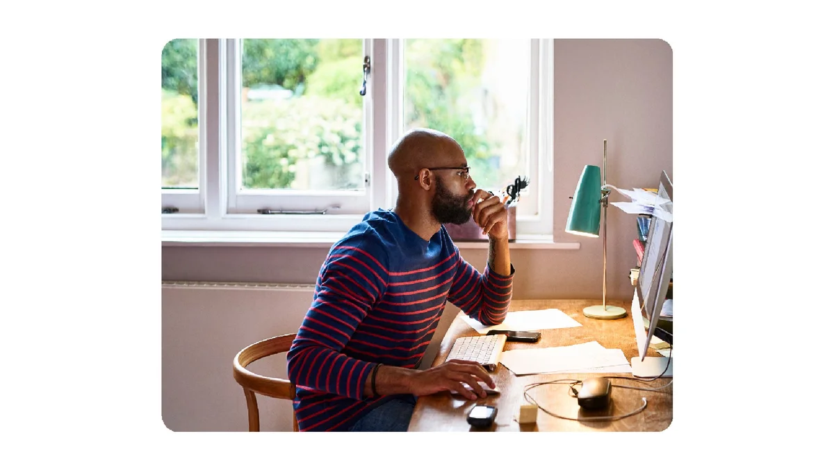 A person sits at a desk next to a window. They are concentrating, looking at a computer desktop screen.