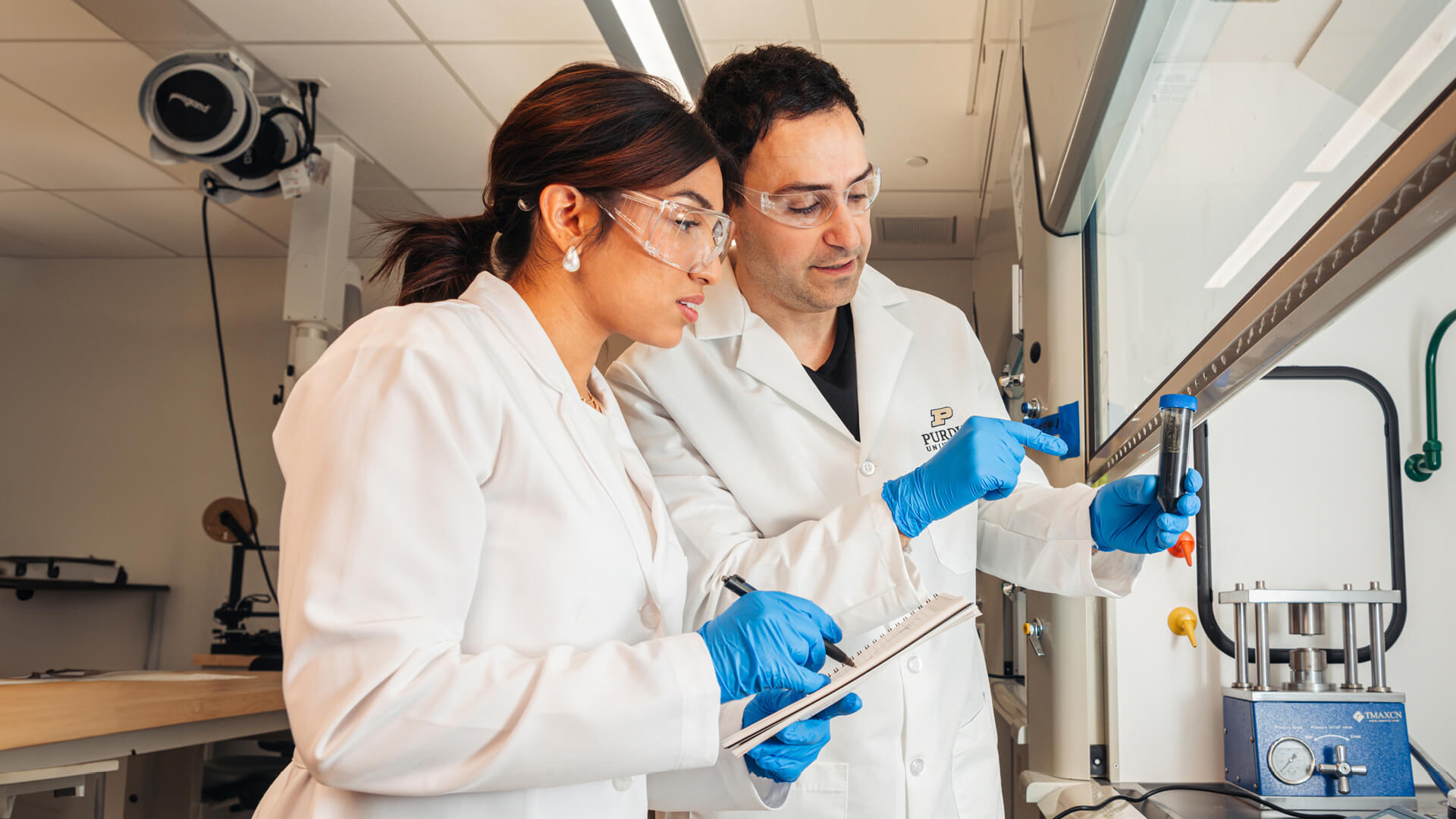 A professor points at a test tube in his hand while a student holding a clipboard looks on.