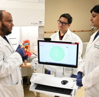 Three people in a laboratory gathered around a 3d bioprinting machine and engaged in discussion 