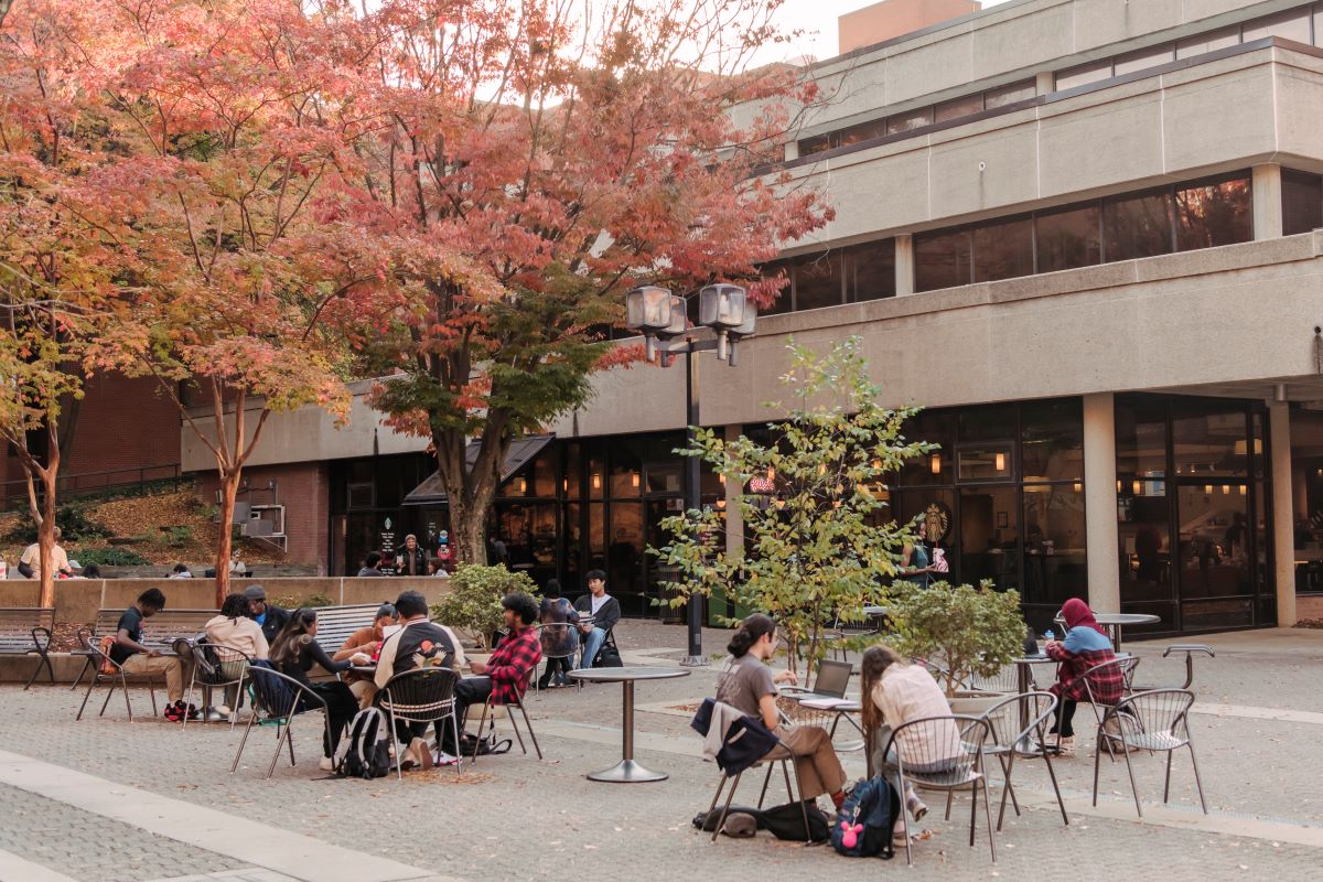 Groups of students sit at tables outside a concrete and glass building.