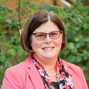 Portrait of Caroline Baker, standing outside in front of greenery, wearing glasses and a pink blazer