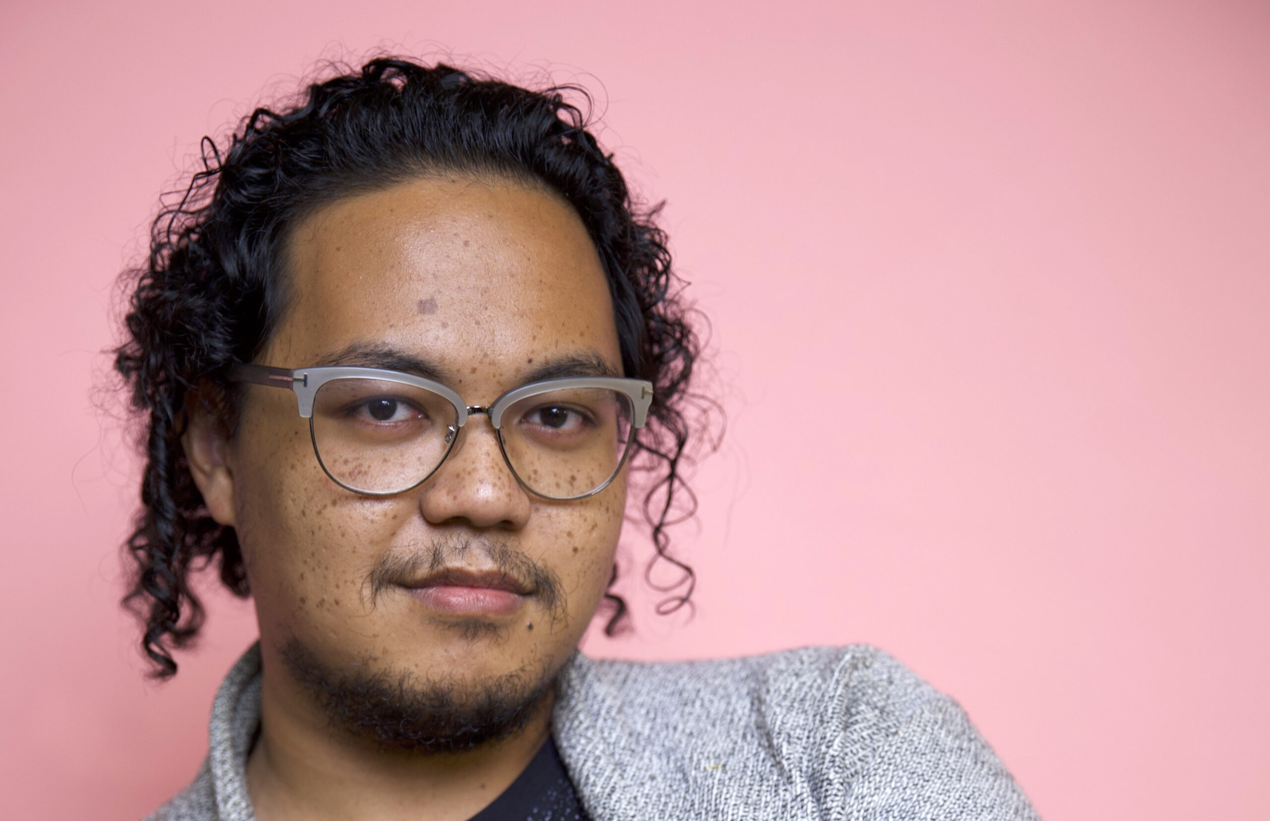 A person with a medium skin tone and dark wavy hair wears glasses and poses against a pink background.