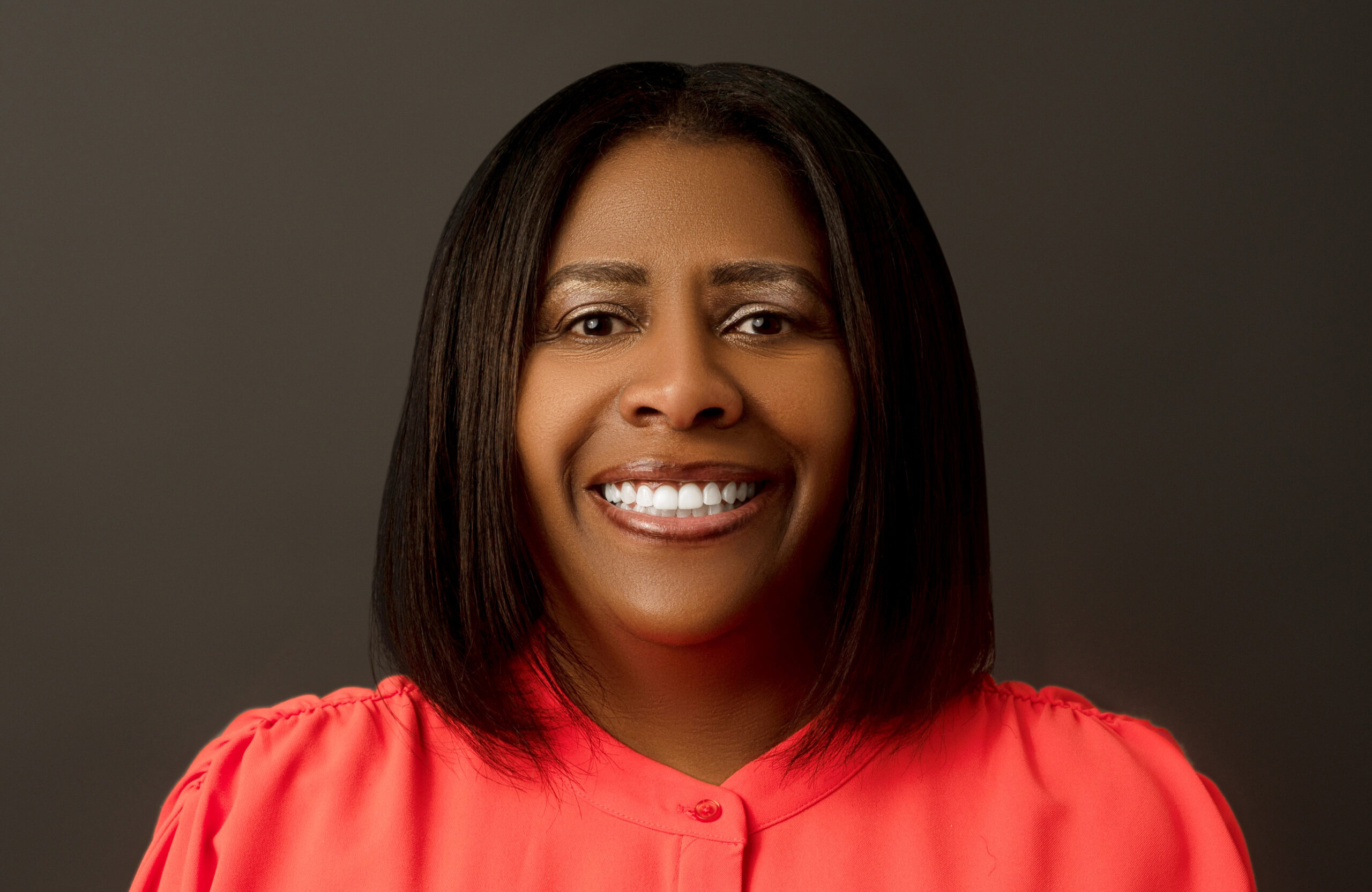 A person with a medium dark skin tone and black hair wears a red top and poses against a brown background.