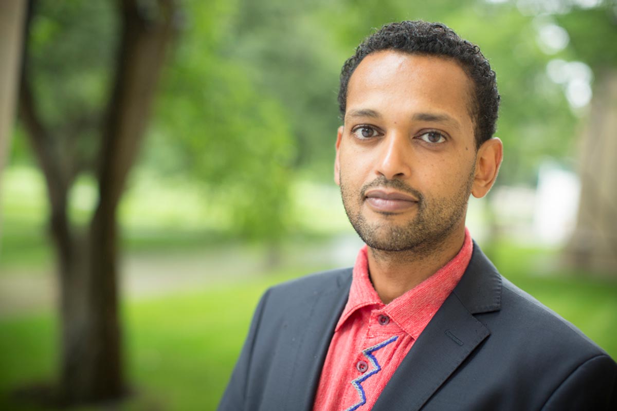 A person with a medium skin tone wearing a red shirt and navy jacket poses against a background of trees and looks at the camera.