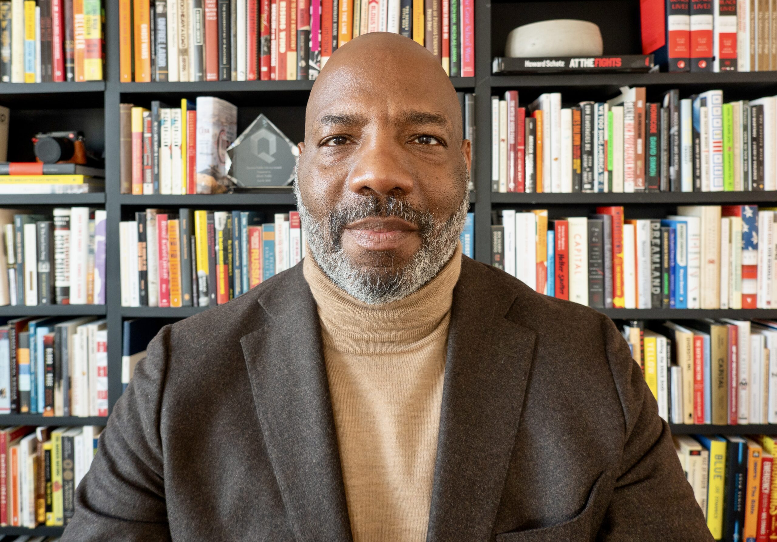 A person with a medium dark skin tone an a short gray beard, wearing a brown jacket, poses in front of shelves of books.