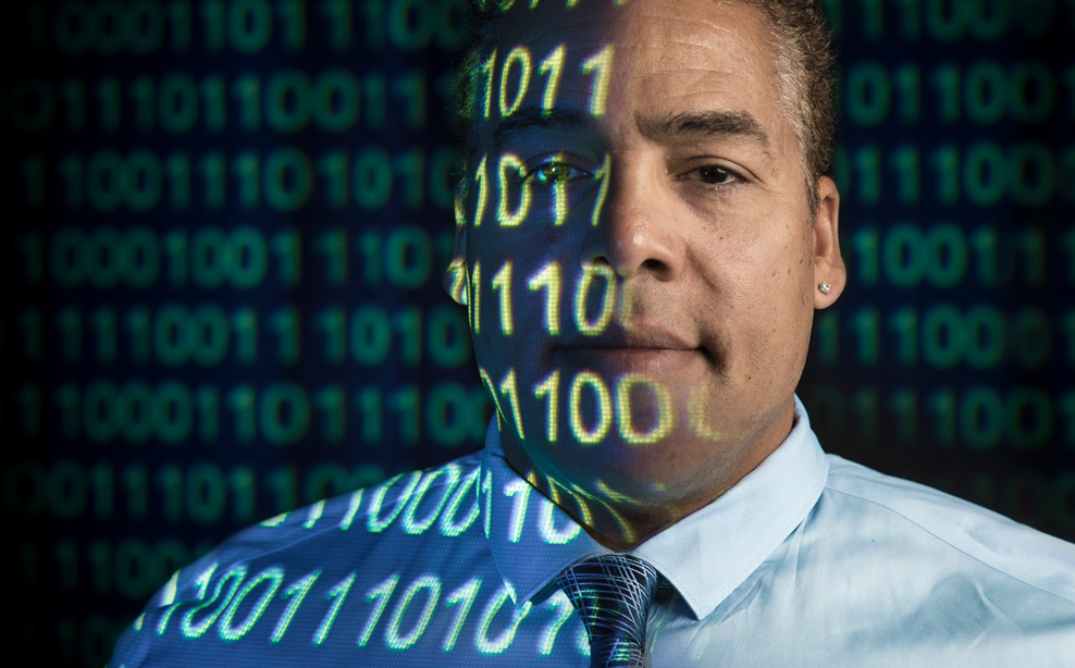 A professor looks at the camera with 1’s and 0’s projected onto his face 